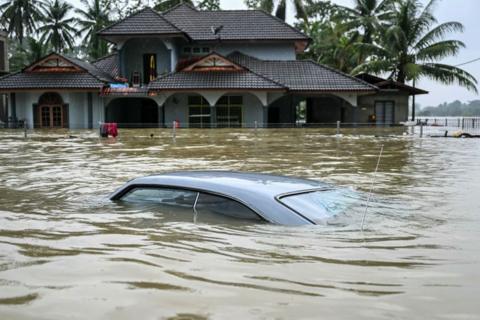 A car submerged almost all the way to the roof in murky floodwater. In the background is a house, also flooded with water at a very high level. 