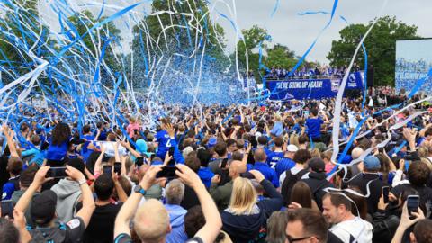 Blue and white streamers and confetti fill the air as fans celebrate the arriving open-top bus of Ipswich Town players and staff