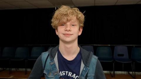 A young boy with curly blonde hair. He is wearing a blue T-shirt and hoody.