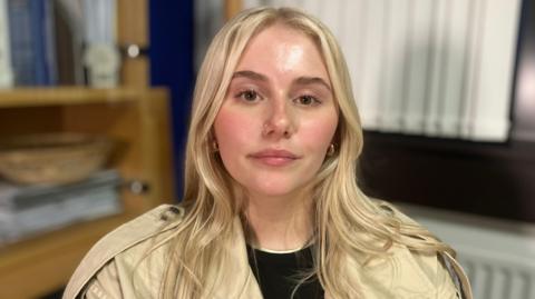 Jayla looking at the camera. She has long blonde hair, brown eyes and a silver hoop nose piercing. She is wearing a cream jacket.
