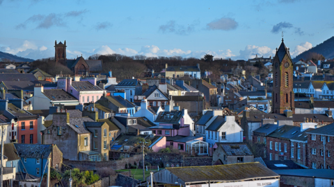 Landscape shot of the village of Peel, Isle of Man. The tops of narrow coastal houses can be seen whilst two church spires are seen in amongst.