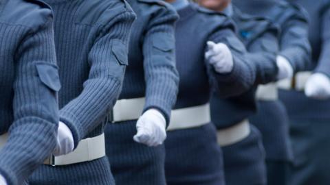 Close-up of people standing in formation wearing blue-grey uniform sweaters, white belts and white gloves, with arms positioned at their sides during a ceremonial parade or drill.