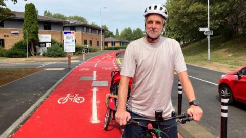 Jim wearing a cream T-shirt and a white helmet on his bicycle- along the red cycle path on Thorpe Wood 