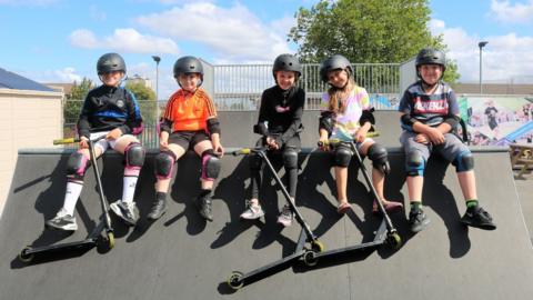 Five children wearing helmets and knee pads sit on top of a ramp at a skate park. Three of them are holding mini scooters.