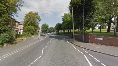 A street view of a road, with trees on both sides of the road. There are red-brick buildings on the left-hand side of the road and a green area to the right. There is a set of traffic lights on the road, and a brick wall on the right-hand side with a street sign saying Peel Street on it.