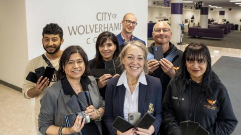 A group of seven people stand in an office holding mobile phones with a City of Wolverhampton Council logo behind them