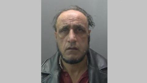 Mugshot of Muhammed Idrees. He is frowning and looking at the camera. He wears a leather jacket with large collars and red shirt underneath. The background is grey. 