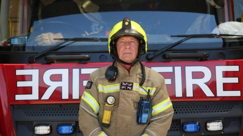 Malcolm McCarthy, a male firefighter standing in front of a bright red fire truck. He is wearing a firefighters uniform - a light brown coat with yellow and silver reflective strips across the chest and upper arms and a bright yellow reflective helmet with a chin strap. The fire truck behind him is seen from the front-on, and has 'Fire' written across the middle