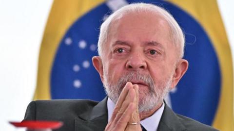 Luiz Inacio Lula da Silva, wearing a grey suit and seated in front of a Brazilian flag, gestures as he attends the approval of a bill to strengthen family farming at the Planalto Palace in Brasilia on 27 November 2024