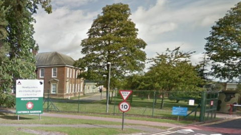 The exterior of a two-storey bricked building with several white windows. There is a green fence around the site and a sign outside which reads 'Headquarters Catterick Garrison'. The site is bordered by trees. 