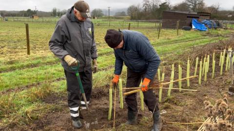 Volunteers at the site in Birling, Kent, planting hedgerows. Both volunteers are in wellingtons and are wearing protective outdoor clothing, including gloves. The volunteer on the left is wearing a cap, with the volunteer on the right in a beanie hat. The volunteer on the right has a number of thin wooden poles in his hand.