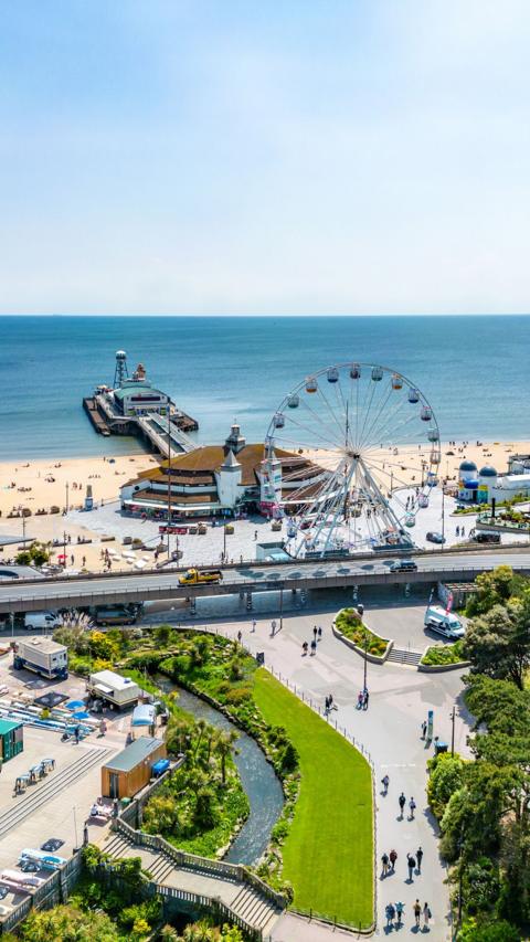 Bournemouth central gardens with the pier, beach and sea behind. The sky is clear blue and its sunny. People are walking along a path next to the grass and a stream. The photo has been taken from an elevated position.
