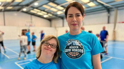 Katherine and Helen standing side-by-side, smiling at the camera. Katherine, who has brown hair, has her arm around Helen who has blonde hair and wears glasses. 