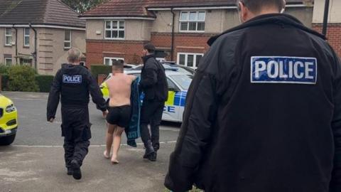 A police officer in a black uniform watches two other police officers handle a man in shorts and no shirt or shoes. He is being led to a police car.