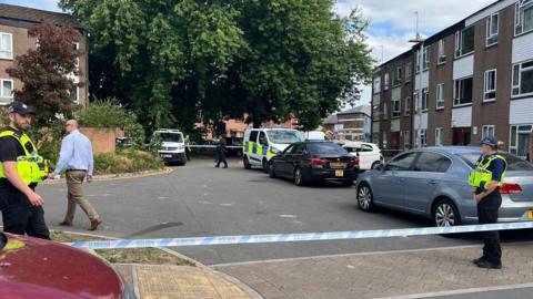 Low rise block of flats with cars parked outside and police van. Two police officers in uniform stand either side of police tape cordoning off the area