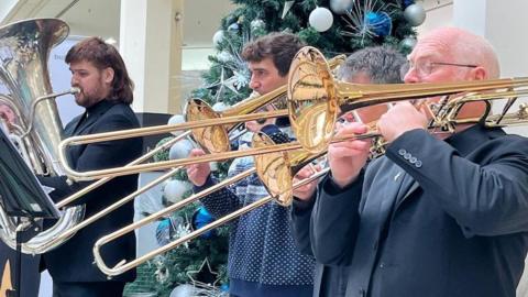 Four men play brass instruments in front of a Christmas tree.