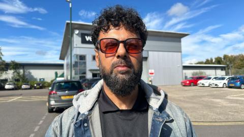 A man standing in a car park with curly black hair and black and grey facial hair. He is wearing red tinted sunglasses, a black t-shirt and a denim jacket.