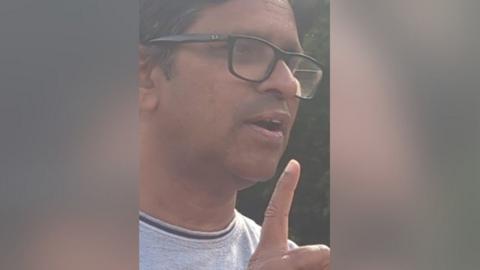 Close up of man's face he has dark, greying hair, is wearing glasses and is mid-speech and holding a finger in the air
