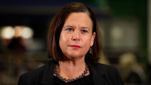 Mary Lou McDonald - a woman with short brown hair wears red lipstick, a necklace with circular stones in the middle, as well as a black suit jacket and a black top. She is looking beyond the camera and the background is blurred.