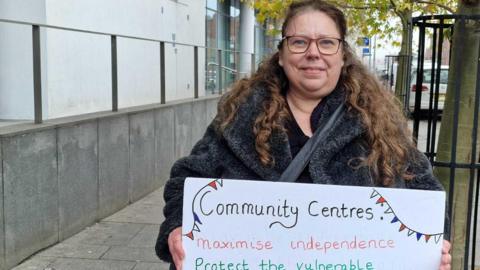 Louise Holland, from the Sherwood Community Centre in Mansfield Road, holding a placard. She is wearing a black furry coat and glasses and is stood outside. 