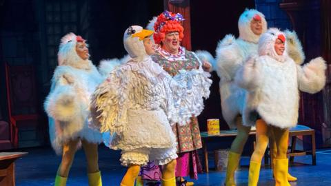 Lucie Goose, played by Dion Davies, is seen in Mother Goose at the Courtyard in Hereford, with ducklings all around her. Lucie Goose has an orange wig and the ducklings all have fluffy white costumes.