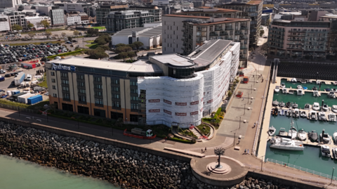 A picture of the front of the Radisson blu in Jersey. It is an all brick building with glass windows and plants in front of it. There is a large glass main entrance in the middle of the image. There is scaffolding surrounding the site.