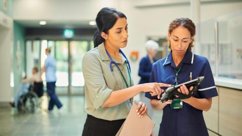 A hospital consultant and senior nurse look at a patient's medical records while standing in a hospital ward.