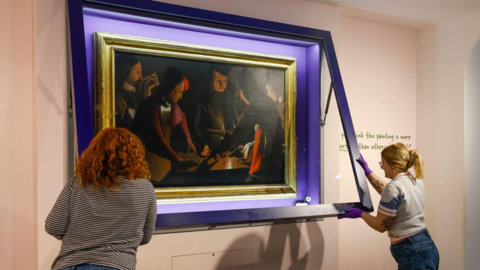 The Dice Players is a painting that features a close-up scene of five people rolling dice on a table. It is quite dark but there is a splash of colour on some of their clothing, including orange and purple. They are wearing formal and elegant clothing. It is inside a large gold frame, which is being placed back into its case on a gallery wall at Preston Park Museum by two members of staff. The two women, pictured from behind, are closing the case.