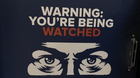 Sign with the words "warning you're being watched" above a drawing of eyes