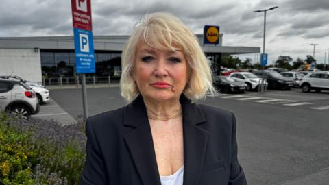 A woman wearing a dark blazer stands beside a parking sign in a Lidl car park. She has blonde hair, lipstick and eyeliner. The supermarket building and several parked cars can be seen in the background. The sign includes the words "new parking rules apply". The sky above is overcast with dark clouds