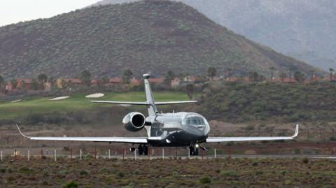 A dark coloured plane taking off on a runway with a green hill in the background. The photo is from the front looking towards the executive-style two-engine aircraft.