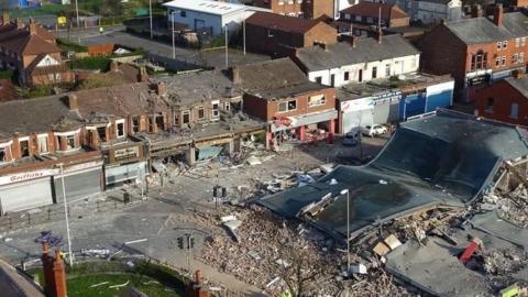An aerial view of New Ferry after the blast showing a row of shops damaged by the explosion and a building completely destroyed. Shop fronts and windows are blown out and the street is full of bricks and debris.