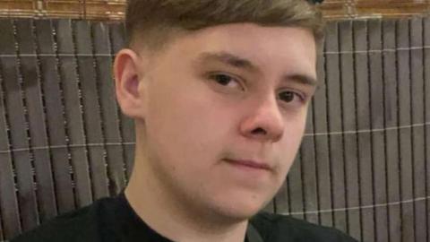 A teenage boy with dark hair. He is starting into the camera. A fence is behind him.