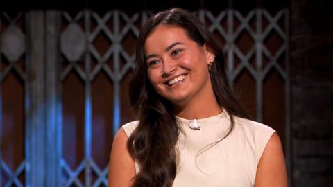 Lucie Macloed of the Hair Syrup company, who has long black hair and is wearing a white dress, smiling on Dragons' Den