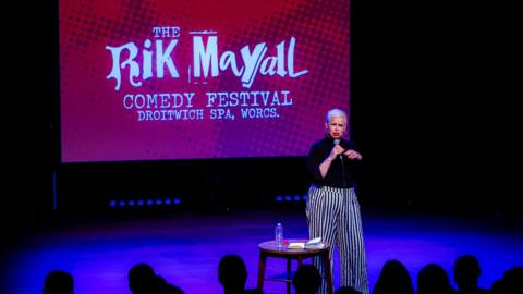 A woman with very short bleach blonde hair and red lipstick is standing on a stage and is speaking into a microphone. She is wearing a black top and black and white striped trousers. Next to her is a small round wooden table with a notebook and water bottle on it. Behind her on a large screen is a red picture with white writing that says "The Rik Mayall comedy festival, Droitwich Spa, Worcs".
