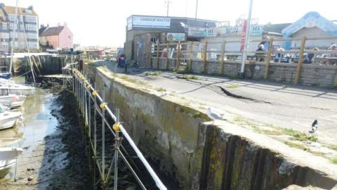The view along the crumbling harbour wall which is lined with scaffolding. The tide is out and several small boats are resting on the mud below. The tarmac path on top of the wall is wobbly with cracks along its length. The side of the concrete wall is also visibly crumbling. On the land side of the path is a low brick wall. Beyond the wall are food outlets and people sitting at picnic benches.