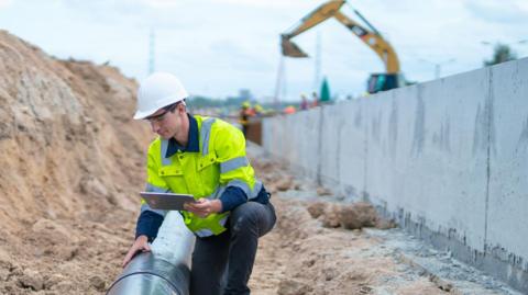 A man in a high-vis jacket, a hard hat and protective goggles leaning over a large pipe holding a tablet in one hand. To the left of frame a pile of dirt, to the right a wall of exposed concrete. In the background and out of focus is a digger.