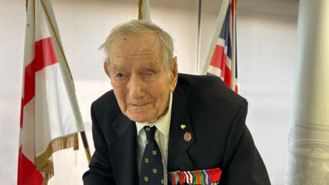 Hundred-year-old Bill Redston wears a dark jacket, white shirt and matching dark tie. He has two pins on his lapel and a series of military medals pinned to his jacket. Behind him are three flags - one of which is the England flag and the other is the union jack.