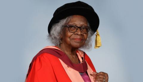 Barbara Dettering wearing her red doctorate robes and holding her honorary graduation scroll, which is white with a red ribbon tied in a bow in the middle. She is wearing a black graduation cap with a gold tassel, and standing against a blue-grey background