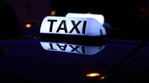 A glowing white taxi sign with bold black lettering sits on the roof of a car at night.