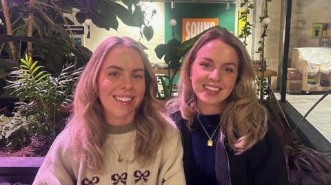 Two women with blonde hair sit in front of some plants. 