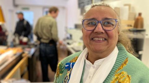 Gulshan Kayembe stands by a cafe counter with people serving in the background