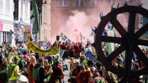 The Hellfire Kernow event in Redruth. The street is filled with people who are wearing costumes and holding banners. There is a wooden wheel in the foreground which has spikes around its and there are clouds of red smoke.