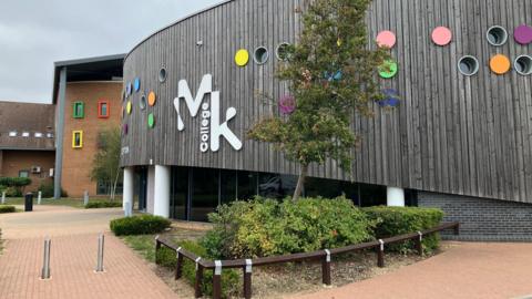 The front of Milton Keynes College, which has a small tree and bushes outside as well as a pedestrianised area. The wooden-clad building has small round windows of different colours.