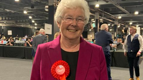 A woman with short white hair wears a pink jacket and a black top and wears a red Labour Party rosette. Behind her are tables and people sat or standing around.