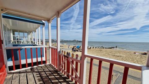 A picture of the beach with the sea and blue sky above taken from the veranda of a beach hut