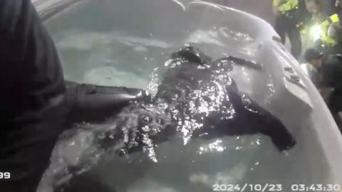 Police in corner of frame arresting a man while black clothing floats in hot tub in the rest of the frame.