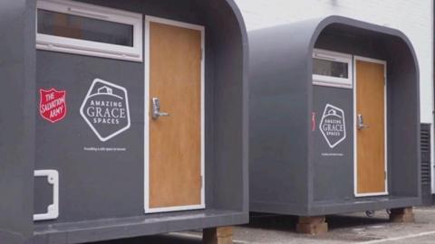 The outside of two grey pods with a small window and brown door with keypad. It has The Salvation Army logo and Amazing Grace Spaces logo side by side.
