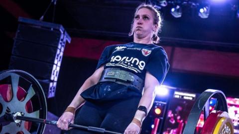 Jade Skill lifts a barbell with various weights on each end. She has dark hair that has been plaited back and wears a black T-shirt with chalk over it. Her cheeks are puffed out with the effort of lifting the weight.