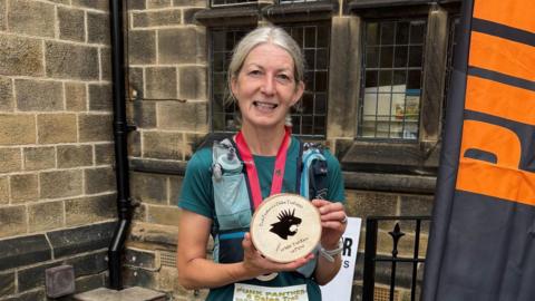 A smiling runner stands outdoors in front of a stone building, holding a circular wooden plaque featuring a black silhouette. She wears a teal athletic top and a race bib. Behind her is a tall banner with the word "PUNK" in bold orange letters.
